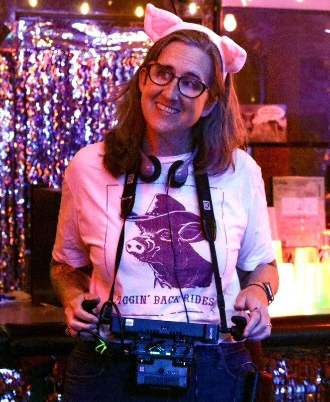 A smiling woman wearing glasses and a pink cat ear headband, holding DJ equipment in a colorful, festive environment with sparkling background and neon lighting.