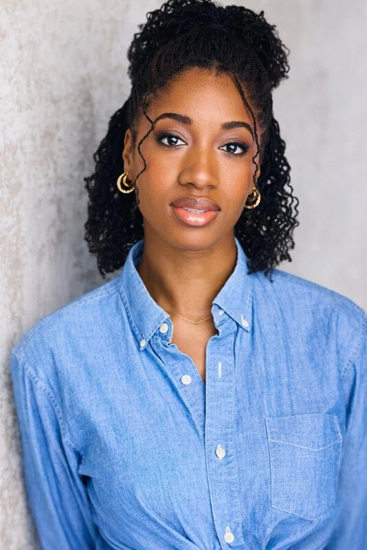 Portrait of a young woman with dark curly hair wearing a light blue denim shirt and gold jewelry, standing against a textured gray wall.