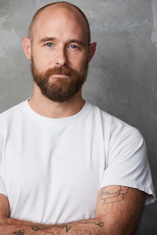 A man with a bald head and beard, wearing a white t-shirt, is standing with his arms crossed against a gray background.