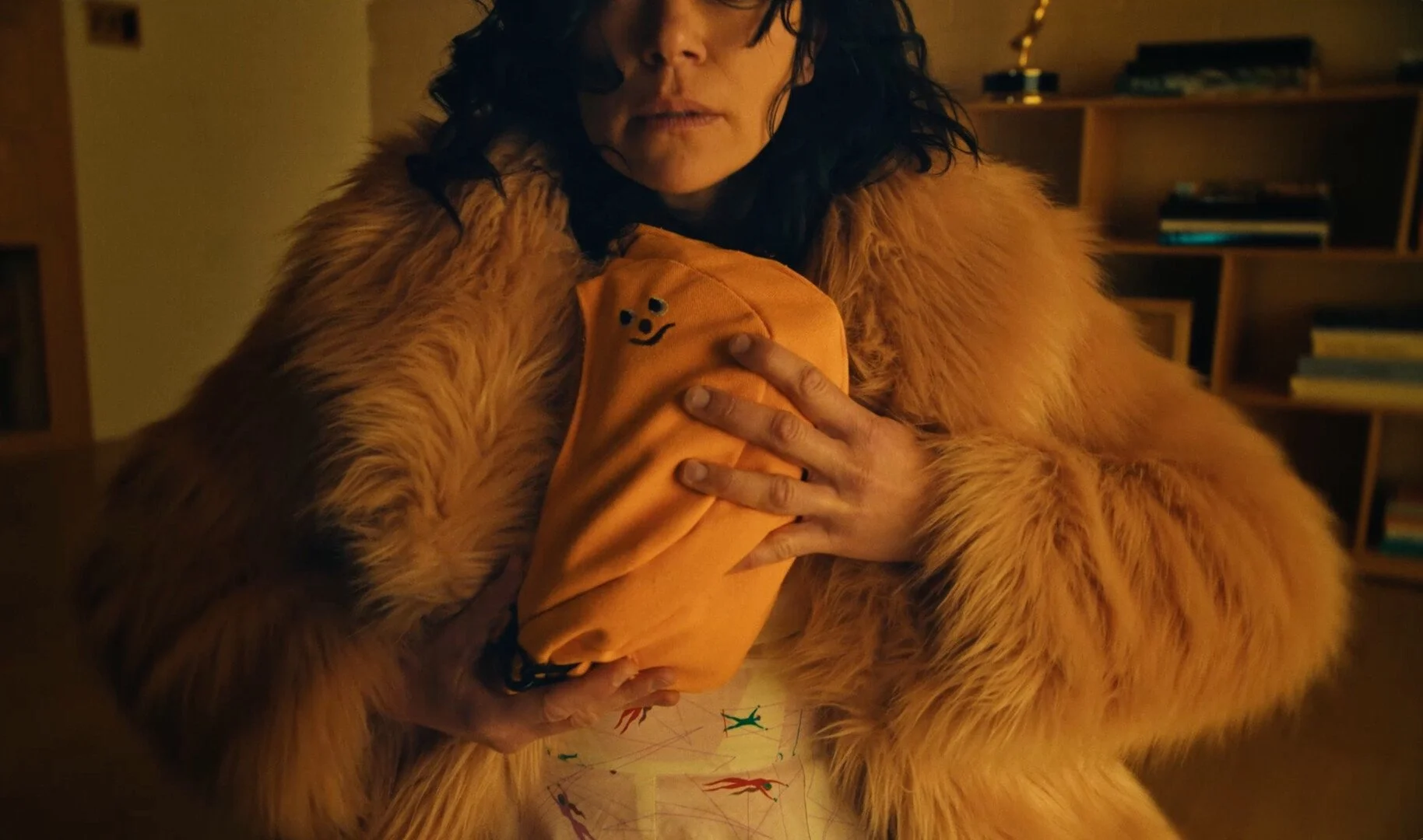 A woman with dark, wavy hair wearing a tan fur coat holds a bright orange fabric with a small smiley face embroidery close to her chest. She is in a warmly-lit room with a bookshelf in the background.