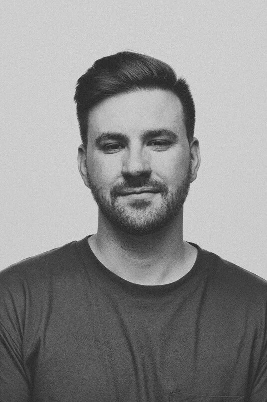 A black and white portrait of a man with styled hair, a beard, and wearing a dark T-shirt, smiling slightly against a plain background.