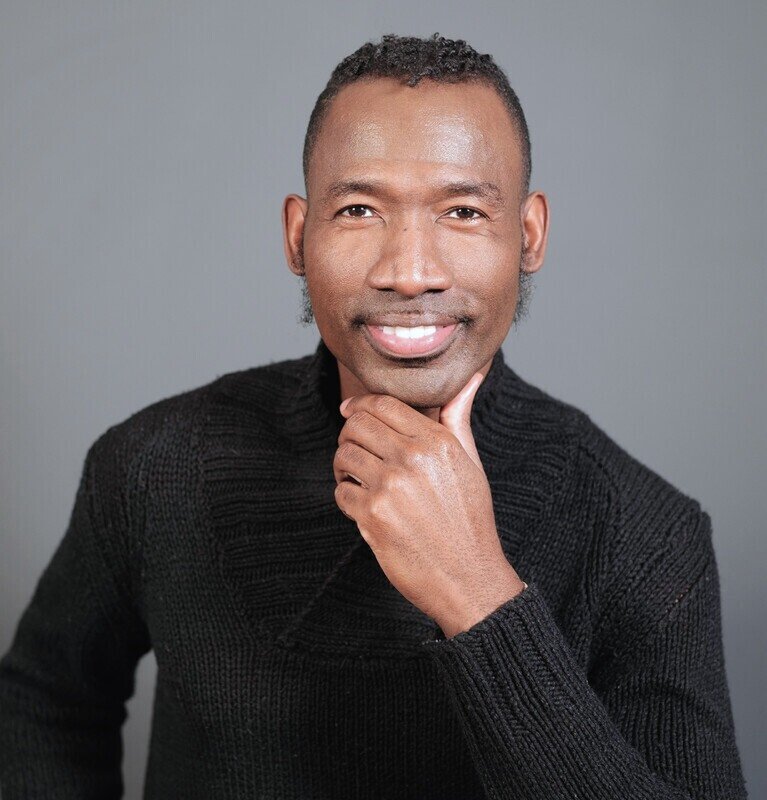 portrait of a man with short curly hair, smiling, wearing a black sweater, hand resting on chin, gray background