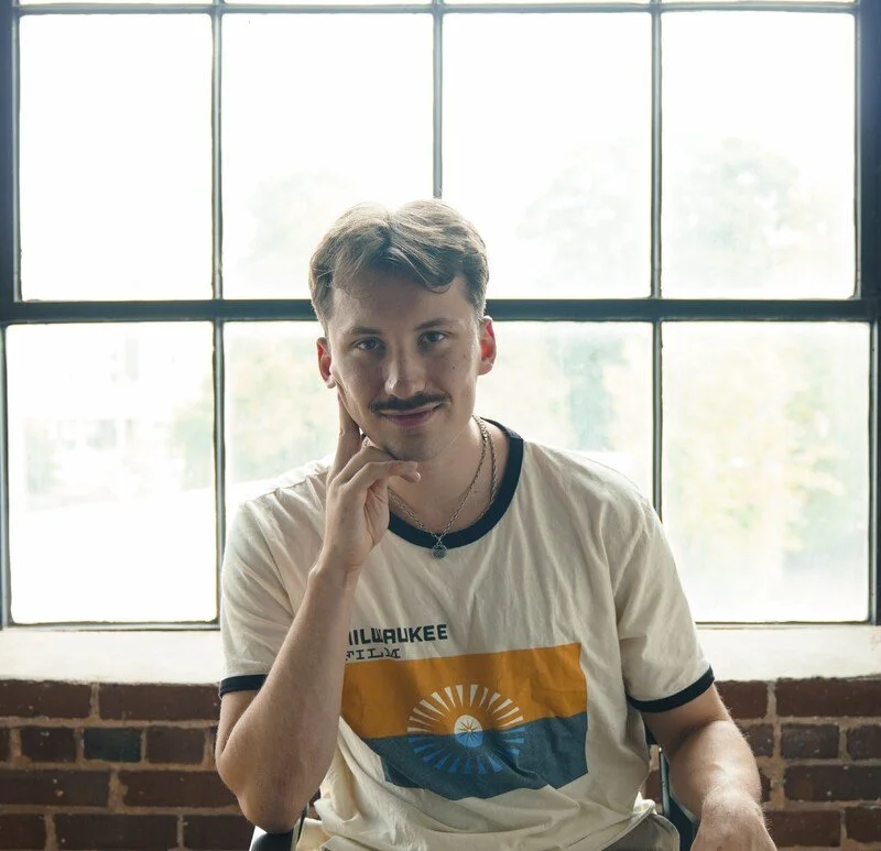 A young man with light brown hair, a mustache, and a goatee sitting in front of a large window with a brick wall below it. He is wearing a white T-shirt with black trim and a colorful graphic featuring the sun and the word "Milwaukee." He is touching his face with his right hand and looking at the camera.