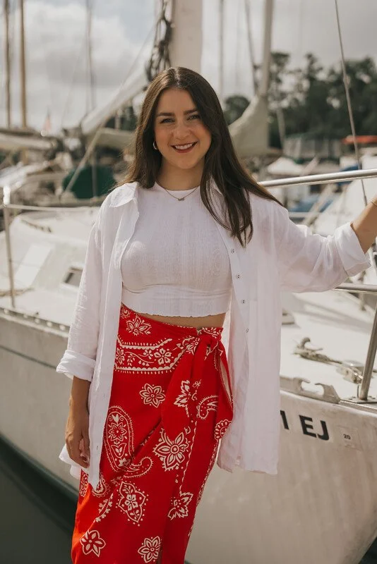 A woman with long dark hair, wearing a white shirt over a cropped top and red bandana pants, standing on a dock next to sailboats.