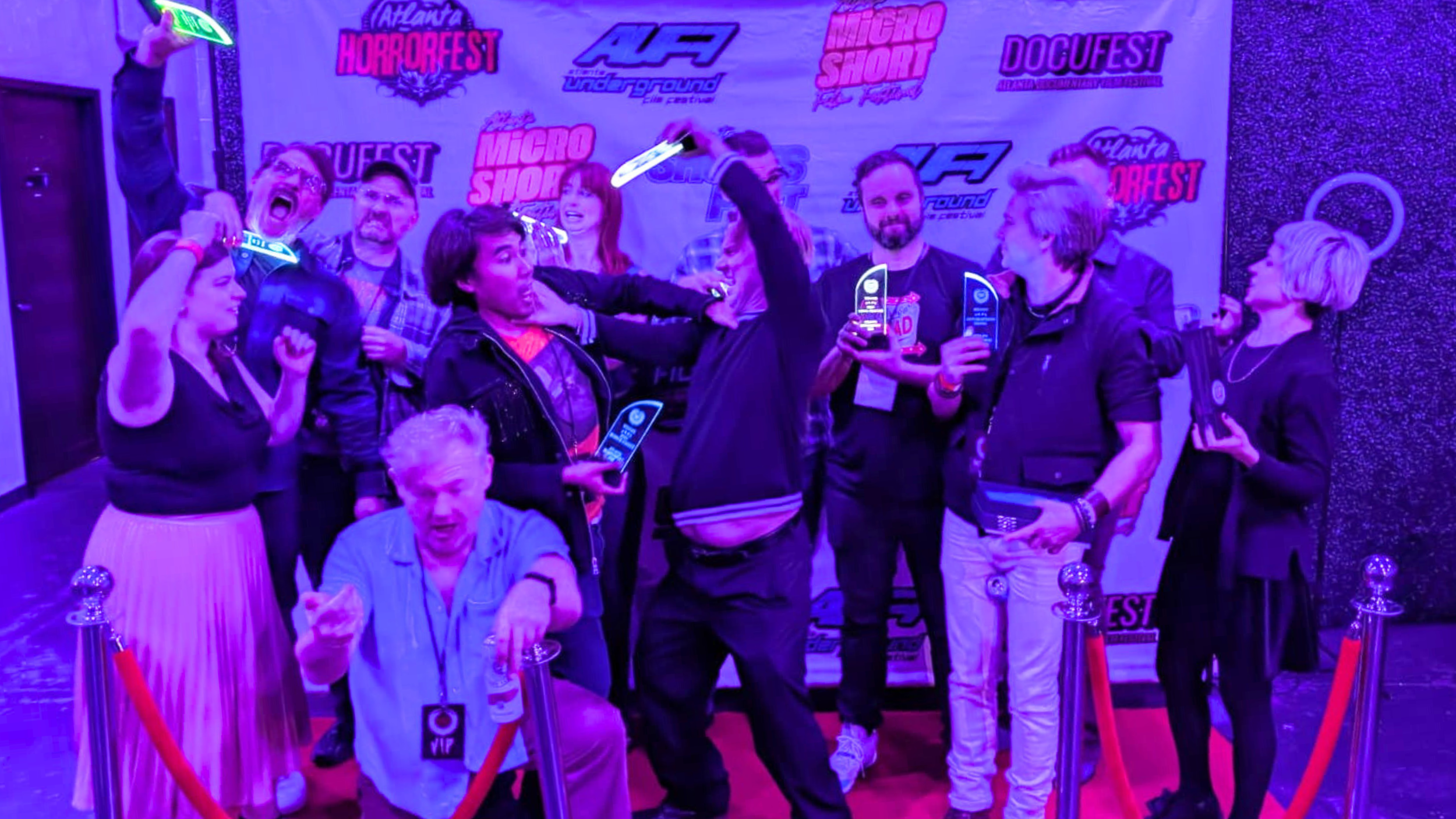 Group of people celebrating at an event, holding awards and posing for a photo in front of a step and repeat banner with logos for Atlanta Horrorfest, Micro Short, and other sponsors. The scene is lit with purple lighting.