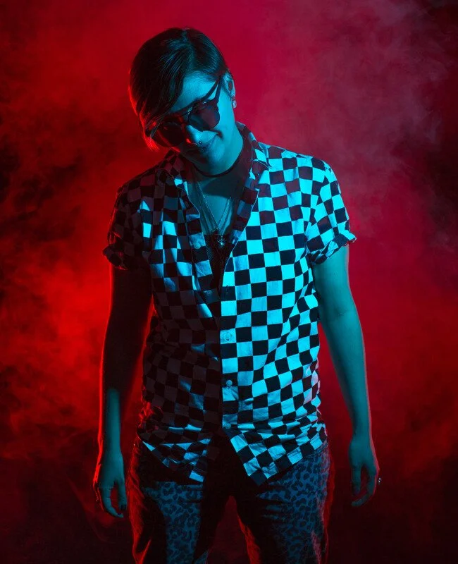 Woman with short hair wearing sunglasses, a checkered button-up shirt, and patterned pants, standing in a smoky environment with red and blue lighting.