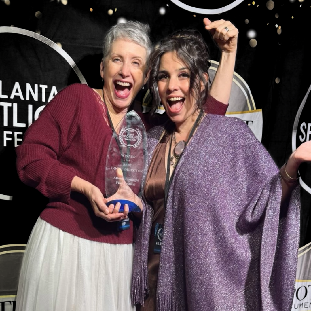 Two women celebrating at an award event, one holding a trophy, both smiling and making jubilant gestures.