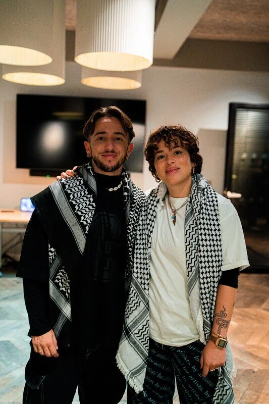 Two young adults standing side by side in a modern indoor setting, both wearing patterned scarves, with smiles on their faces.