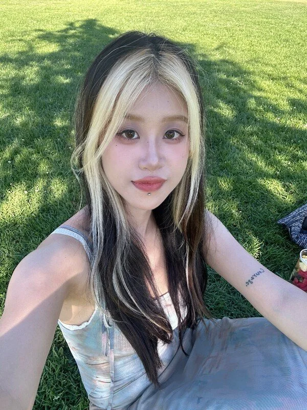 Young woman with long black and blonde hair sitting on grass in park, taking selfie.