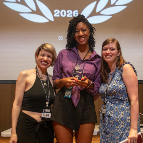 Three women smiling on stage during an award ceremony, with a large screen behind them displaying a laurel wreath and the year 2026.