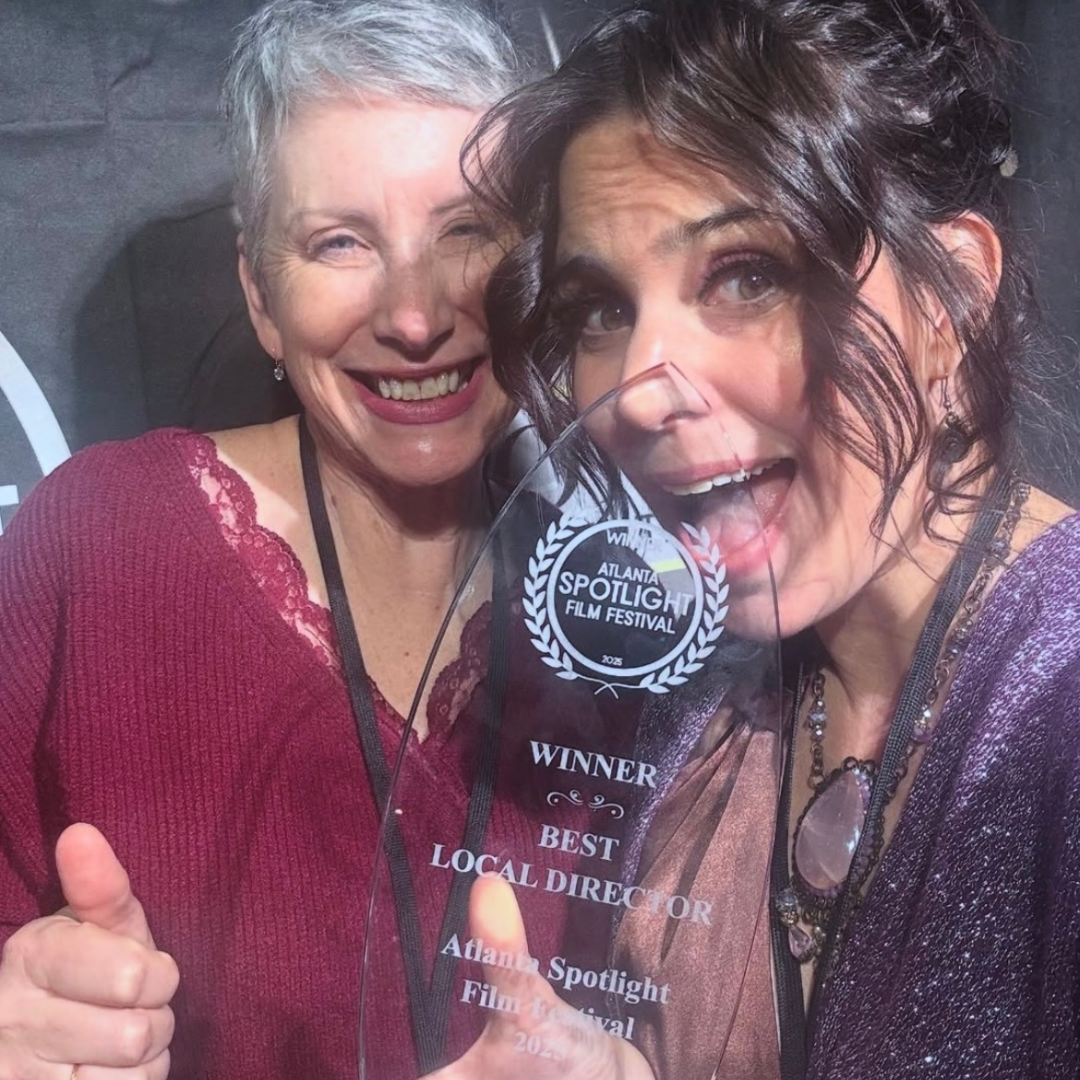 Two women smiling and holding an award trophy at the Atlanta Spotlight Film Festival, with one giving a thumbs-up and the other showing excitement.