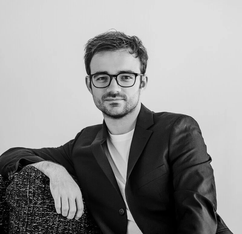 Black and white portrait of a young man with glasses, wearing a blazer over a t-shirt, sitting and leaning on a textured armrest with a plain background.