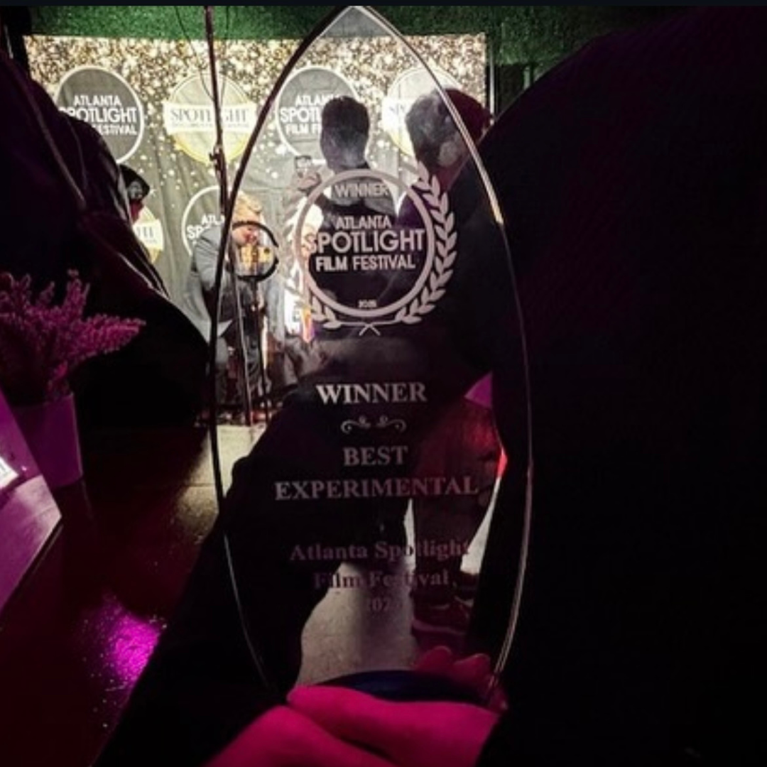 An award glass trophy with inscriptions indicating it was won at the Atlanta Spotlight Film Festival for Best Experimental film. The background shows people taking photos and a decorated backdrop with festival logos.