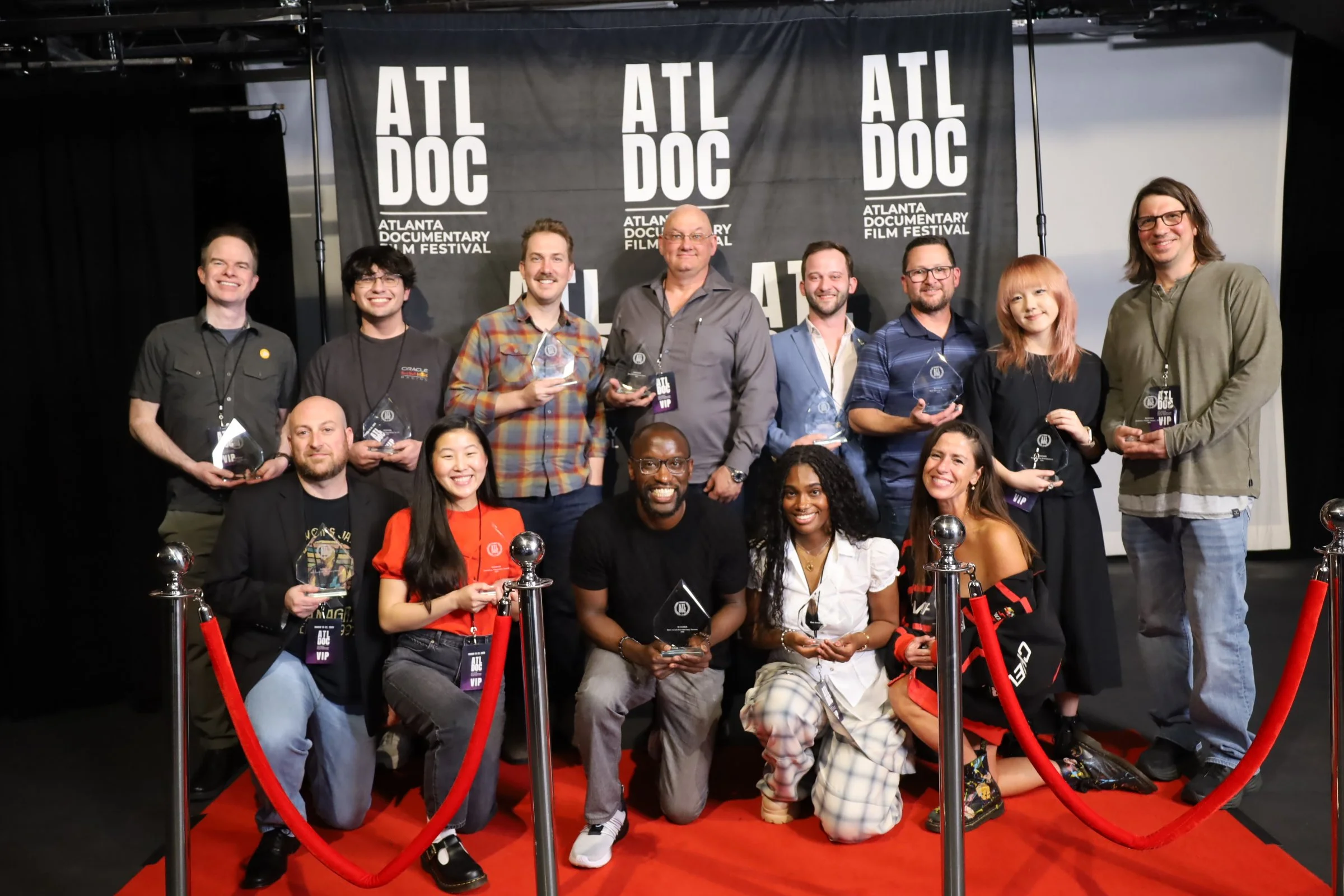 A group of people is gathered on a stage at the Atlanta Documentary Film Festival, holding awards, with some smiling and posing for the camera. Behind them is a black backdrop with the festival's logo and text, and red velvet ropes surround the stage.