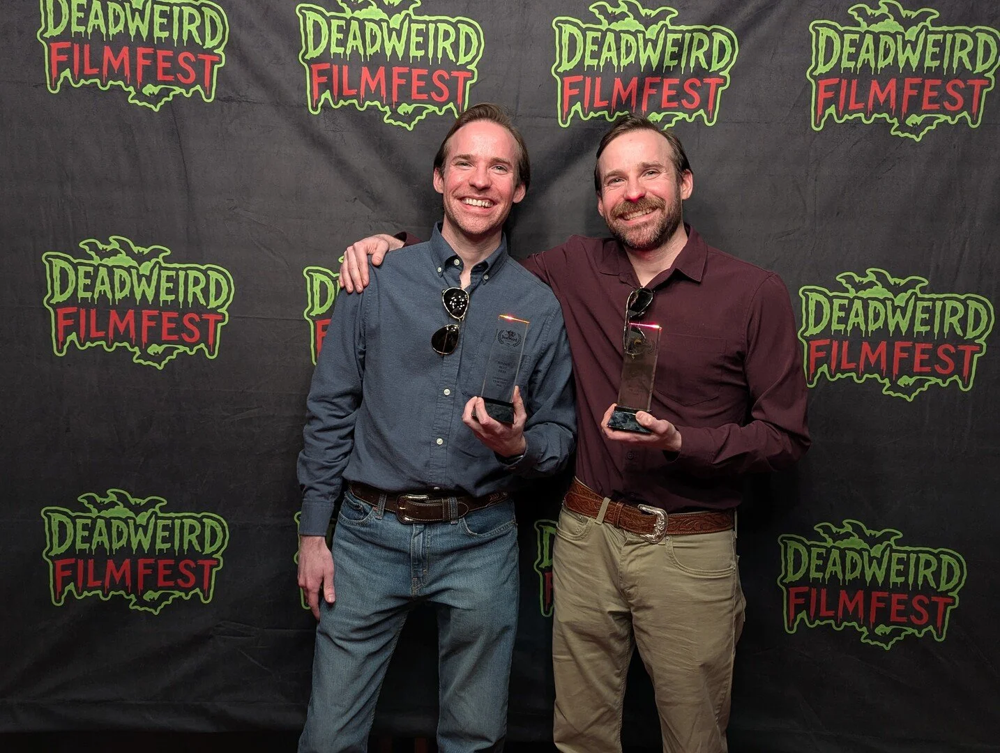 Two smiling men standing in front of a Halloween-themed backdrop with the logo "Deadweird Film Fest" repeated, holding awards, one with sunglasses hanging from his shirt collar, their arms around each other's shoulders.
