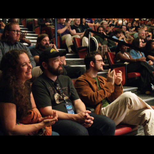 Audience attending an event in a theater or auditorium, seated in rows, some clapping and smiling.