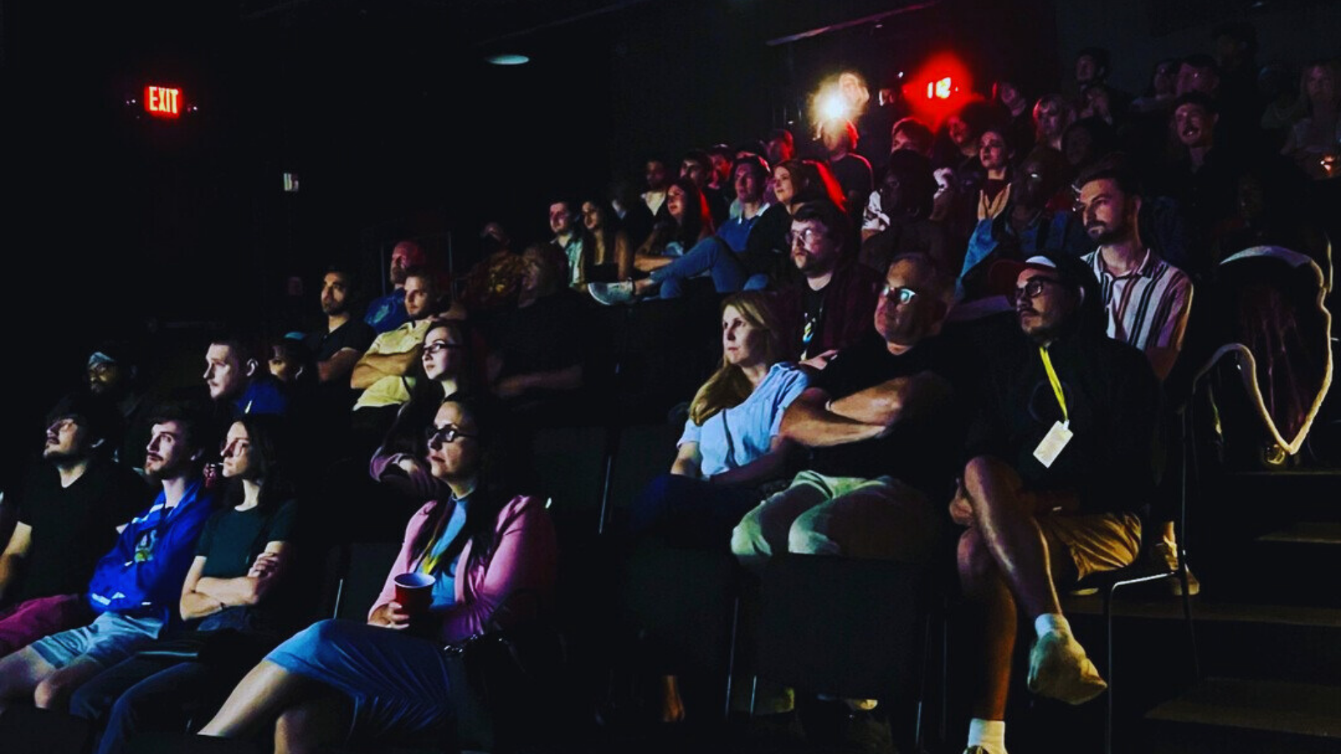 Audience members sitting in a dark theater watching a performance or film with focused expressions, illuminated by dim light.