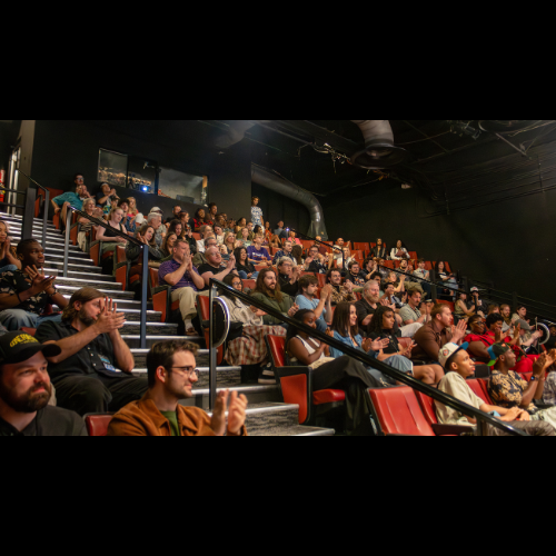 Audience members seated in a theater, watching and clapping during a performance or presentation.