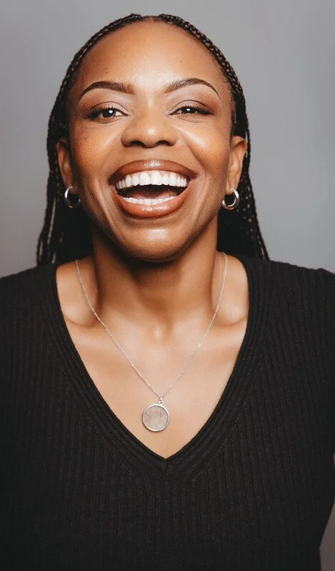 A woman smiling with her tongue out, wearing a black top, hoop earrings, and a necklace with a round pendant, against a gray background.