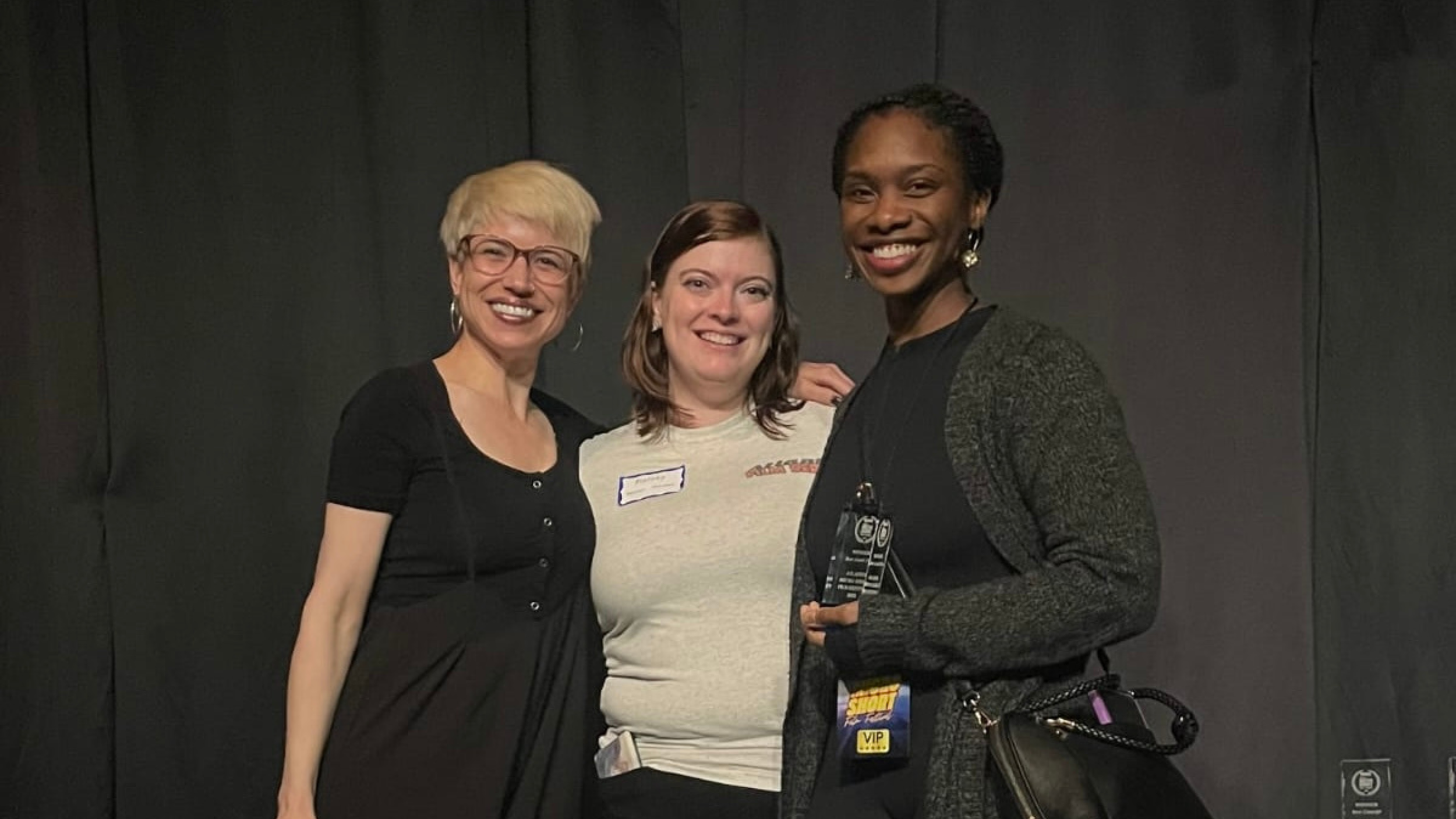 Three women standing together, smiling, in front of a dark wall. One woman on the right holds an award and has a VIP badge. The woman on the far left has short blonde hair and glasses. The woman in the middle has shoulder-length brown hair. The woman on the right has dark, styled hair and earrings.