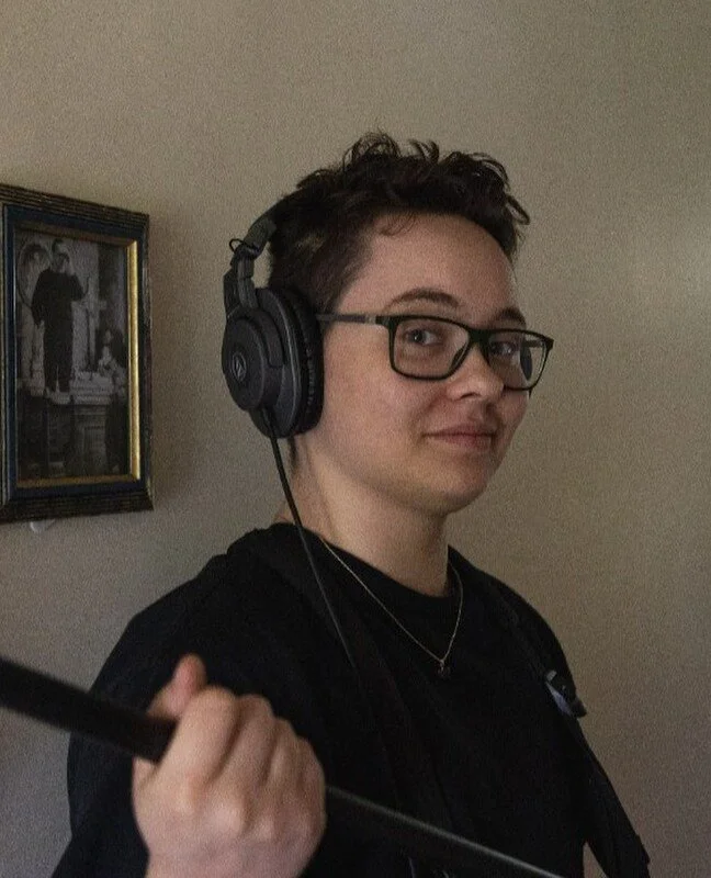 A young woman with short dark hair, glasses, and a slight smile, wearing over-ear headphones and a black shirt, holding what appears to be a broom or mop handle, standing indoors with a wall-mounted framed black-and-white photograph in the background.