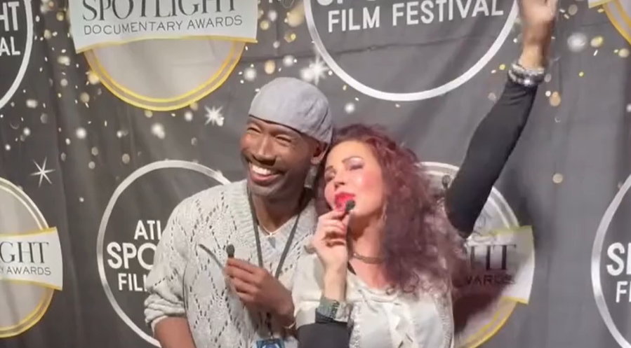 Two people, a man and a woman, smiling and posing for a photo at an event with a backdrop that reads 'Spotlight Documentary Awards' and 'Atlanta Film Festival'. The woman is holding a microphone and raising her arm, while the man is holding a small object and smiling.
