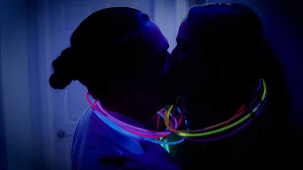 Two women are kissing in a dark room, wearing glow-in-the-dark necklaces.