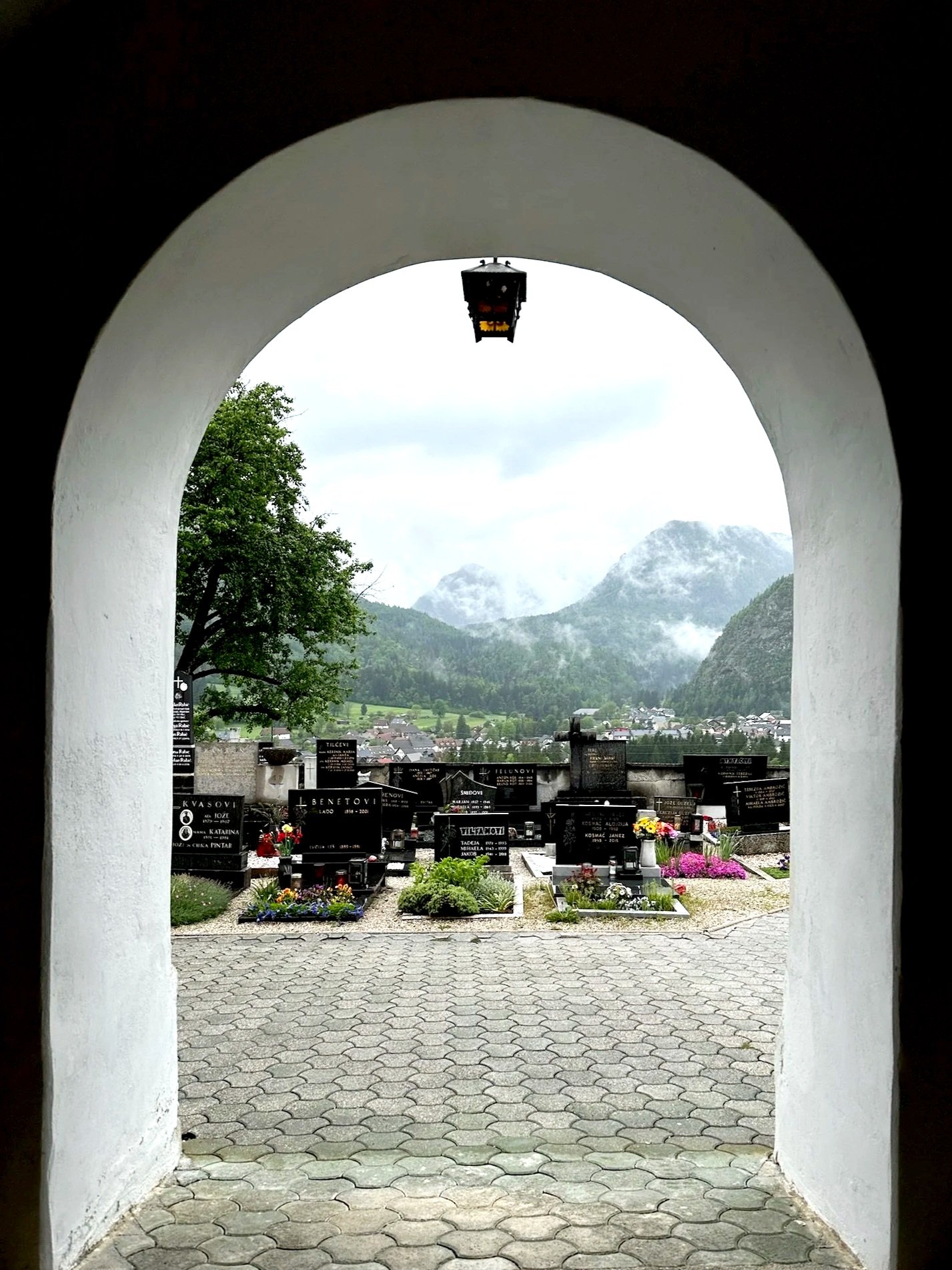 View of a cemetery with tombstones and flowers, seen through an arched window, with mountains and a cloudy sky in the background.