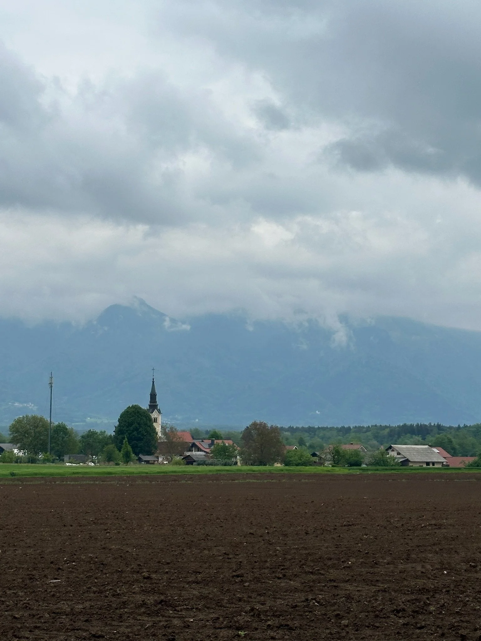 A rural landscape with freshly plowed dark soil in the foreground, green fields with trees and small houses in the middle ground, and a distant church with a tall spire, under a cloudy sky with mountain ranges in the background.