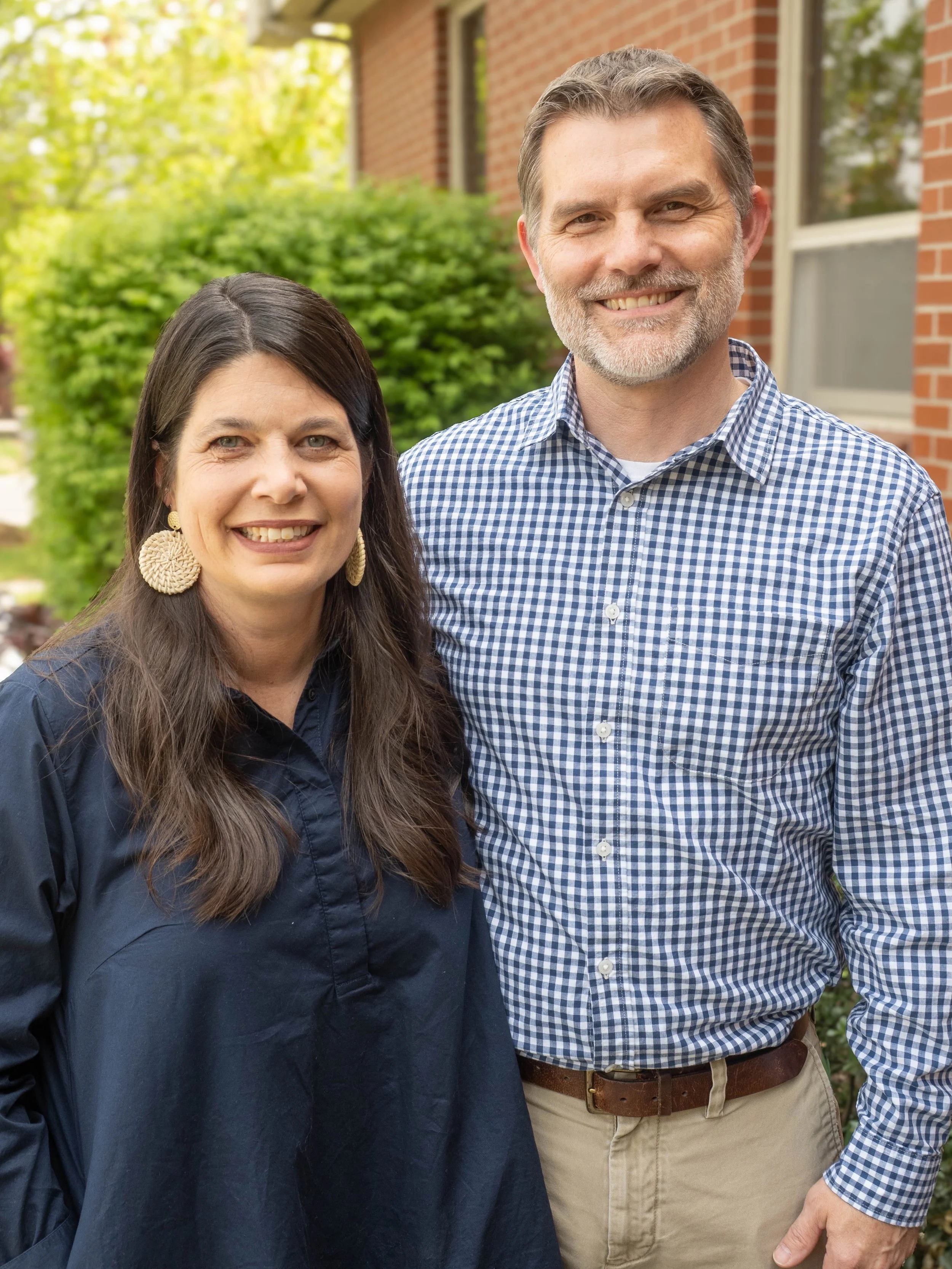 A smiling middle-aged woman with long dark hair and large woven earrings stands next to a smiling middle-aged man with short gray hair and beard. They are outdoors in front of a brick building with green shrubbery and trees in the background.