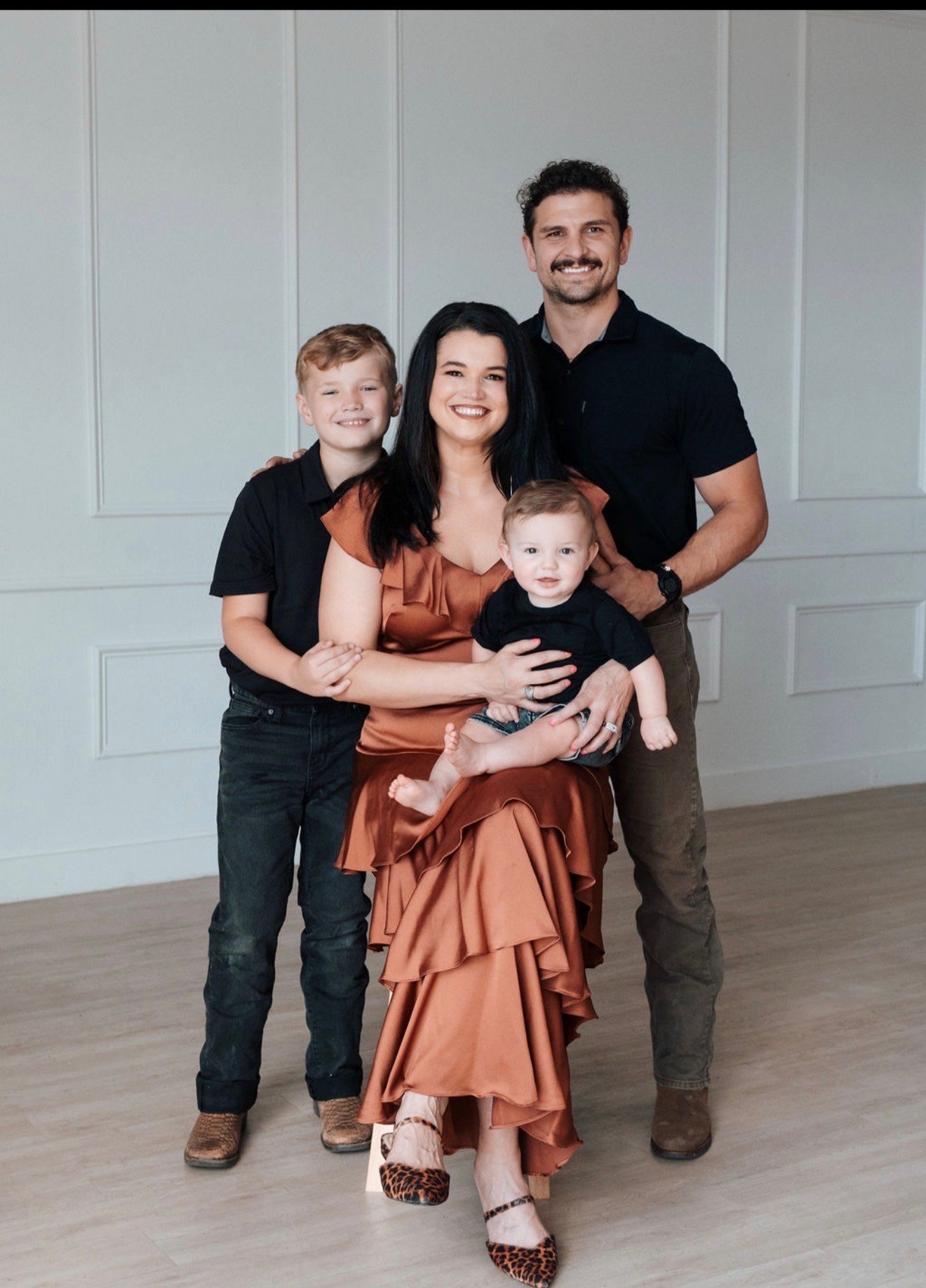 A family of five posing in front of a white paneled wall. The woman, dressed in an orange satin dress, is seated and holding a young child on her lap. Two boys and a man stand behind her, smiling. The children are wearing black shirts, and the man is in a black polo. The woman is wearing leopard print shoes.