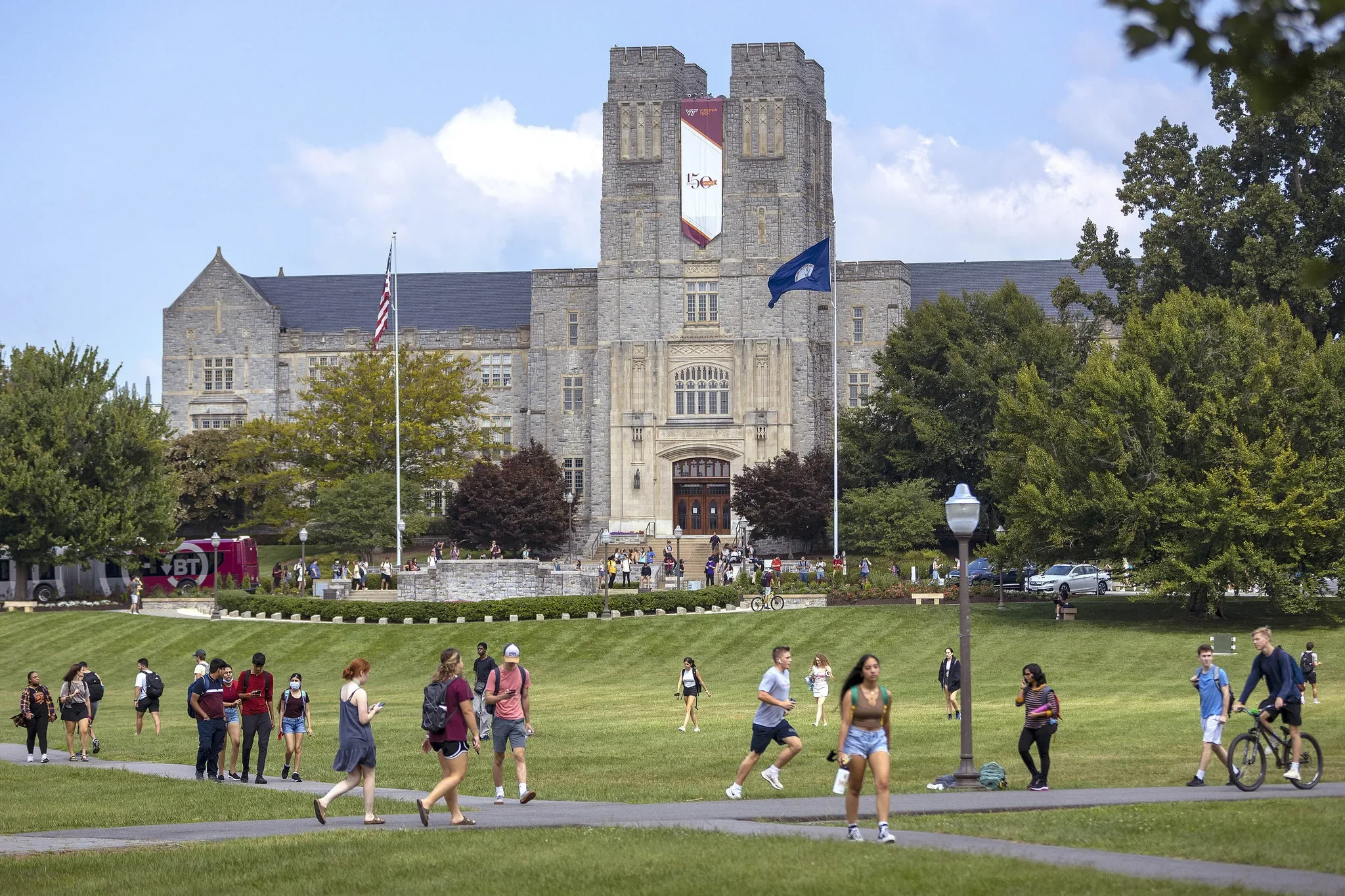 A university campus with a stone castle-like building in the background, surrounded by green trees and a crowd of students walking, sitting, and biking in the grass and pathways.