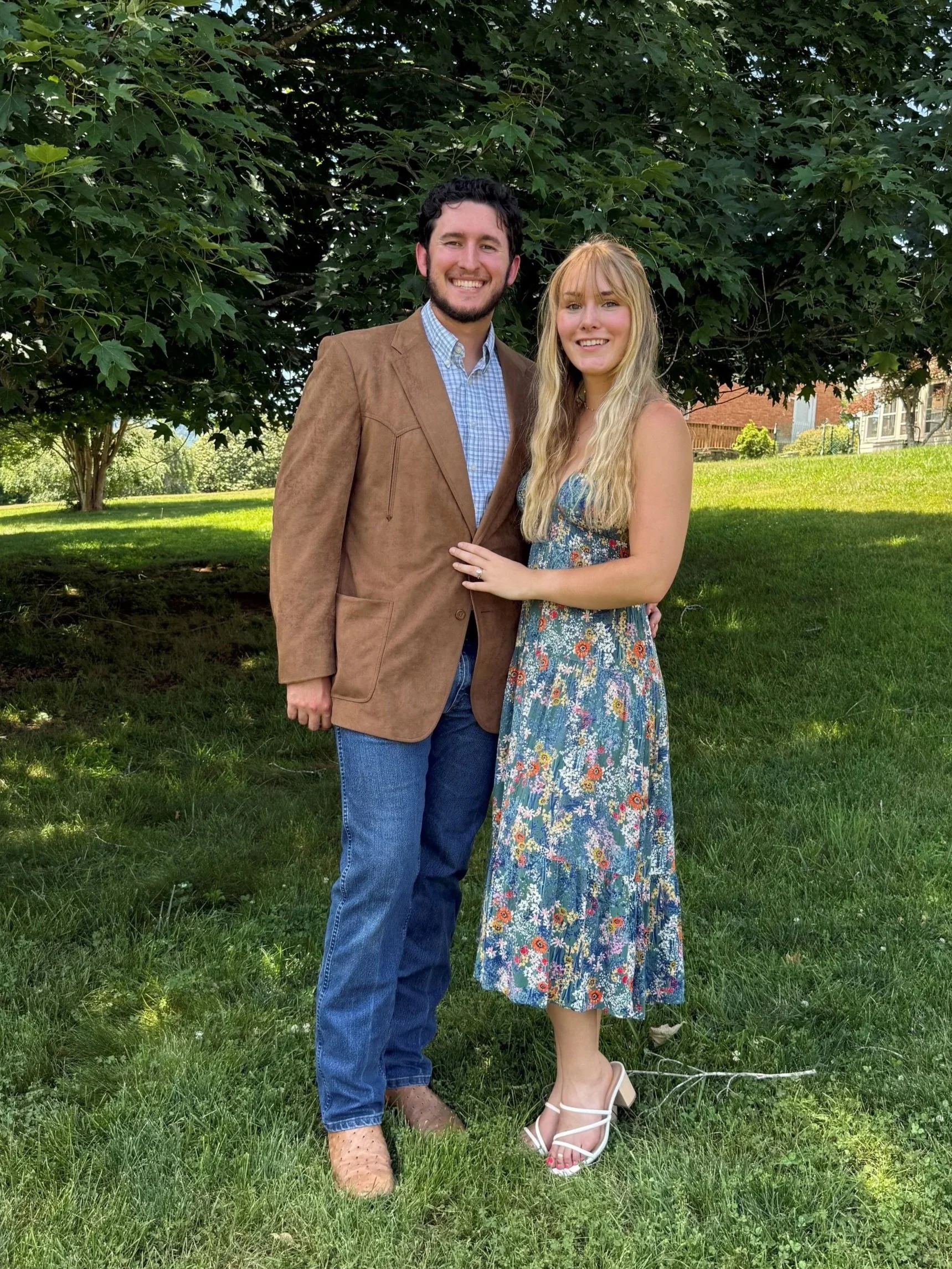 A smiling man in a tan blazer and blue jeans standing next to a smiling woman in a floral dress and white heels, outdoors on grass with trees and a house in the background.