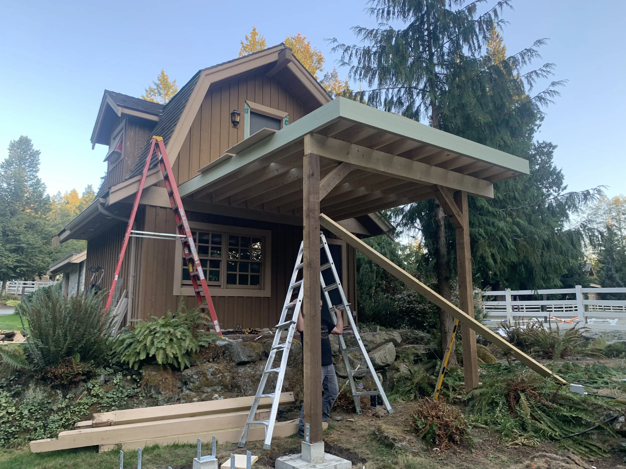 A house under construction with a deck extension being built. There are ladders, construction tools, and workers on-site, surrounded by trees and a white fence in the background.