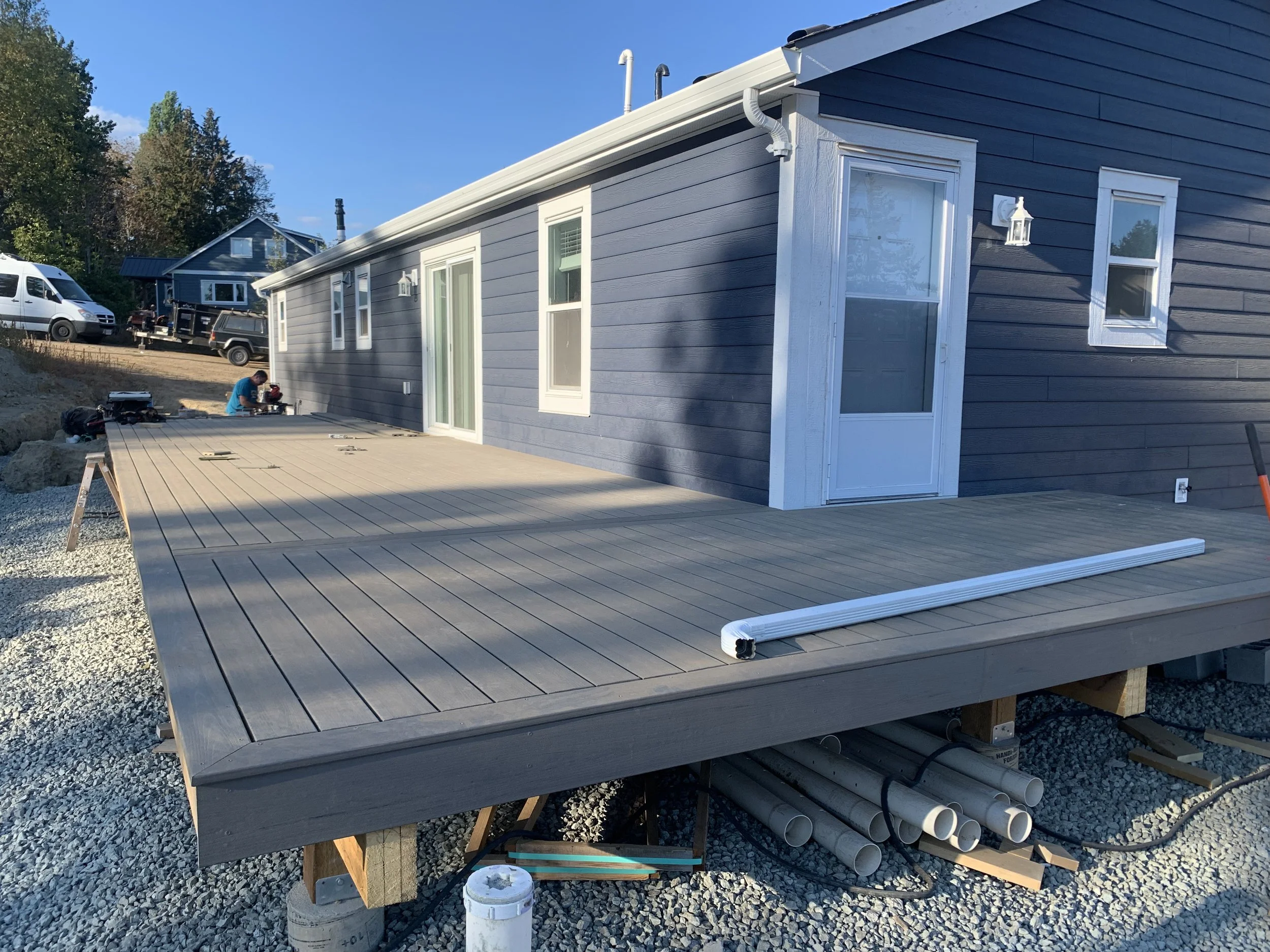 Building a house with gray siding and a wooden deck, construction workers installing the deck, with construction materials and equipment on site.