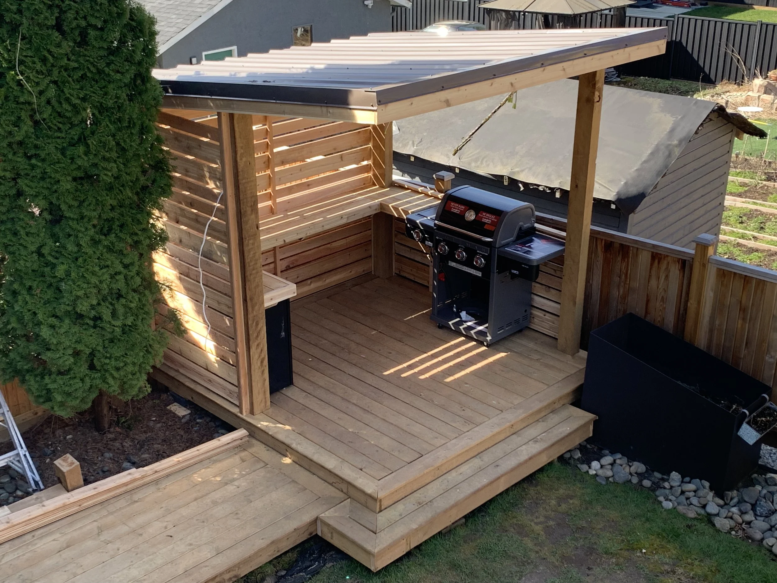 A wooden backyard deck under construction with a black gas grill on it. The deck has a partially completed roof with a metal canopy and horizontal wooden slats on the side. There is a large green tree on the left, and a garden with grass and plants below.