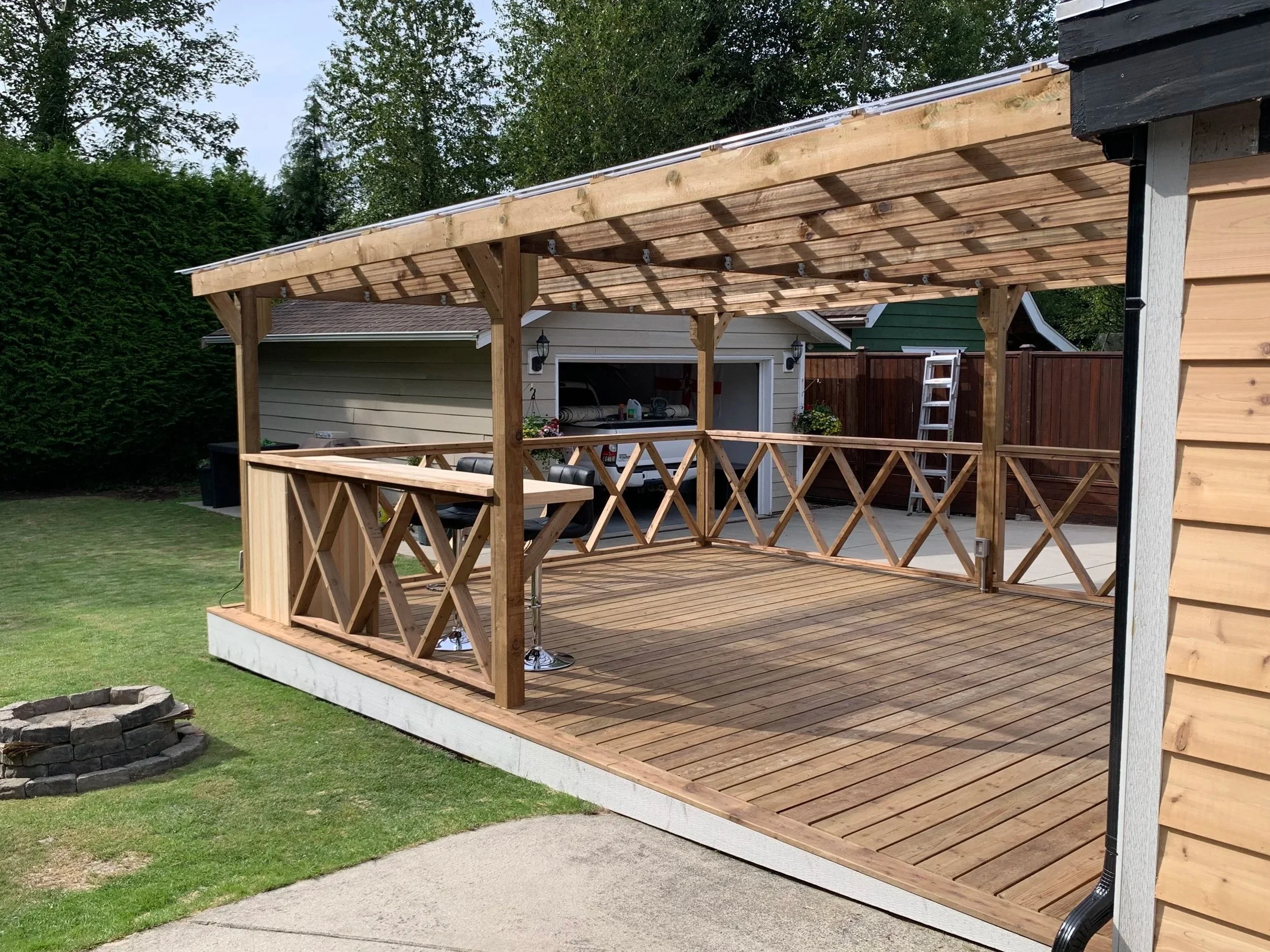 A backyard deck with a wooden railing and posts, constructed with natural wood, attached to a house with beige siding, with a house and garage visible in the background. There are trees and grass surrounding the area.