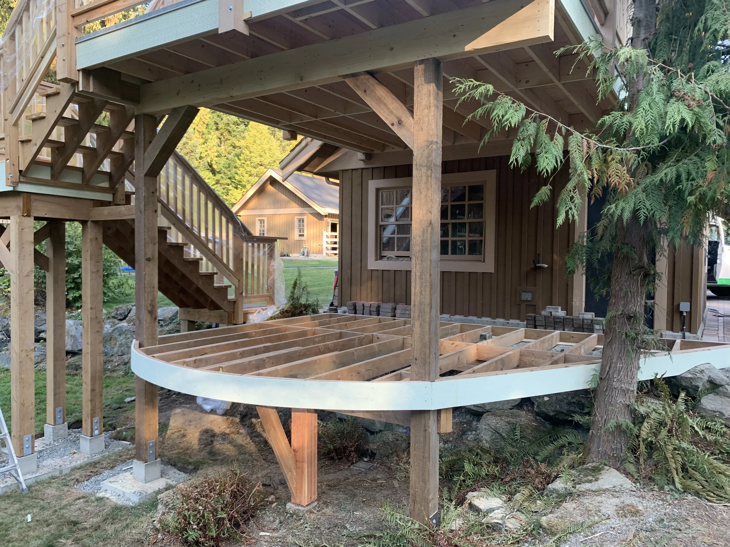 A wooden deck under construction attached to a house, with stairs leading up to it. The structure is supported by posts, and some flooring and railing are installed. There are trees and other houses in the background.