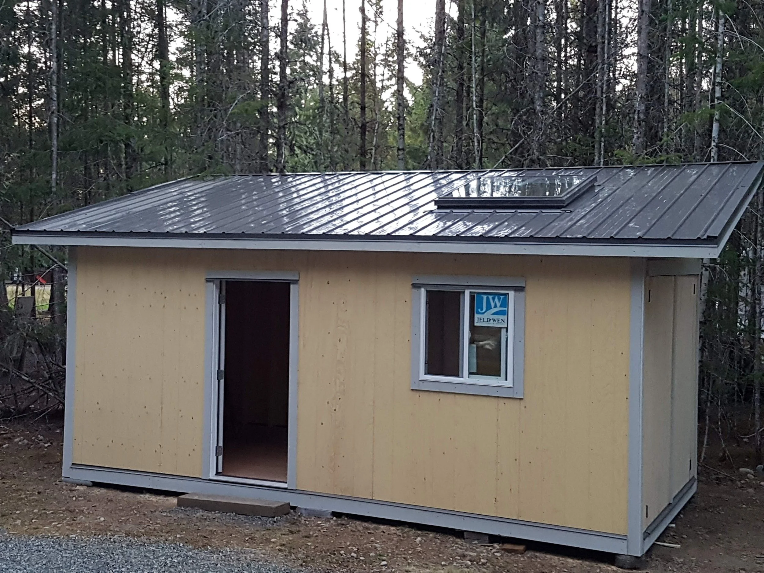 A small yellow shed with a metal roof and a skylight window, situated in a wooded area with trees in the background.