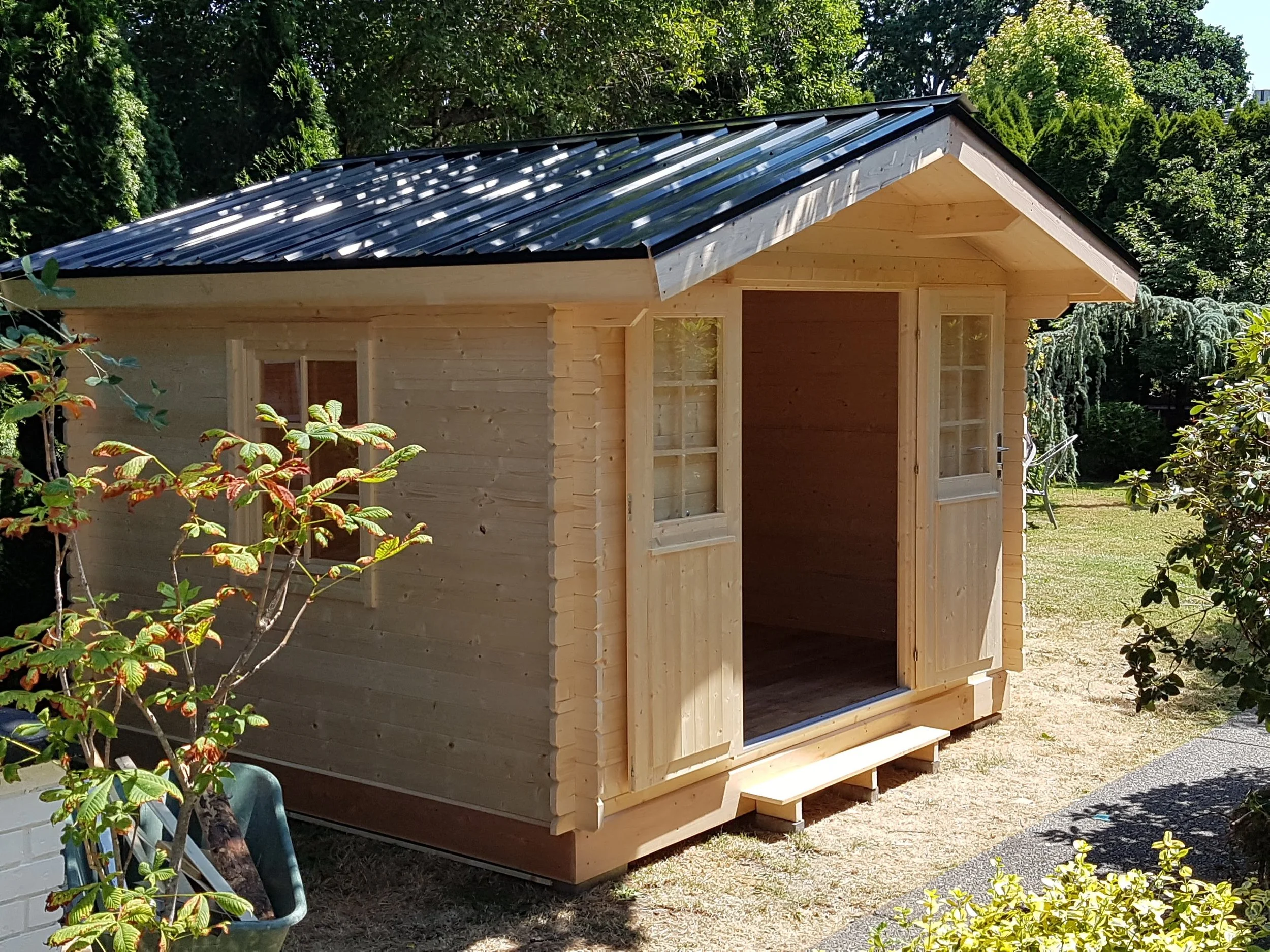 A small unfinished wooden shed with a black metal roof, situated outdoors among trees and shrubs.