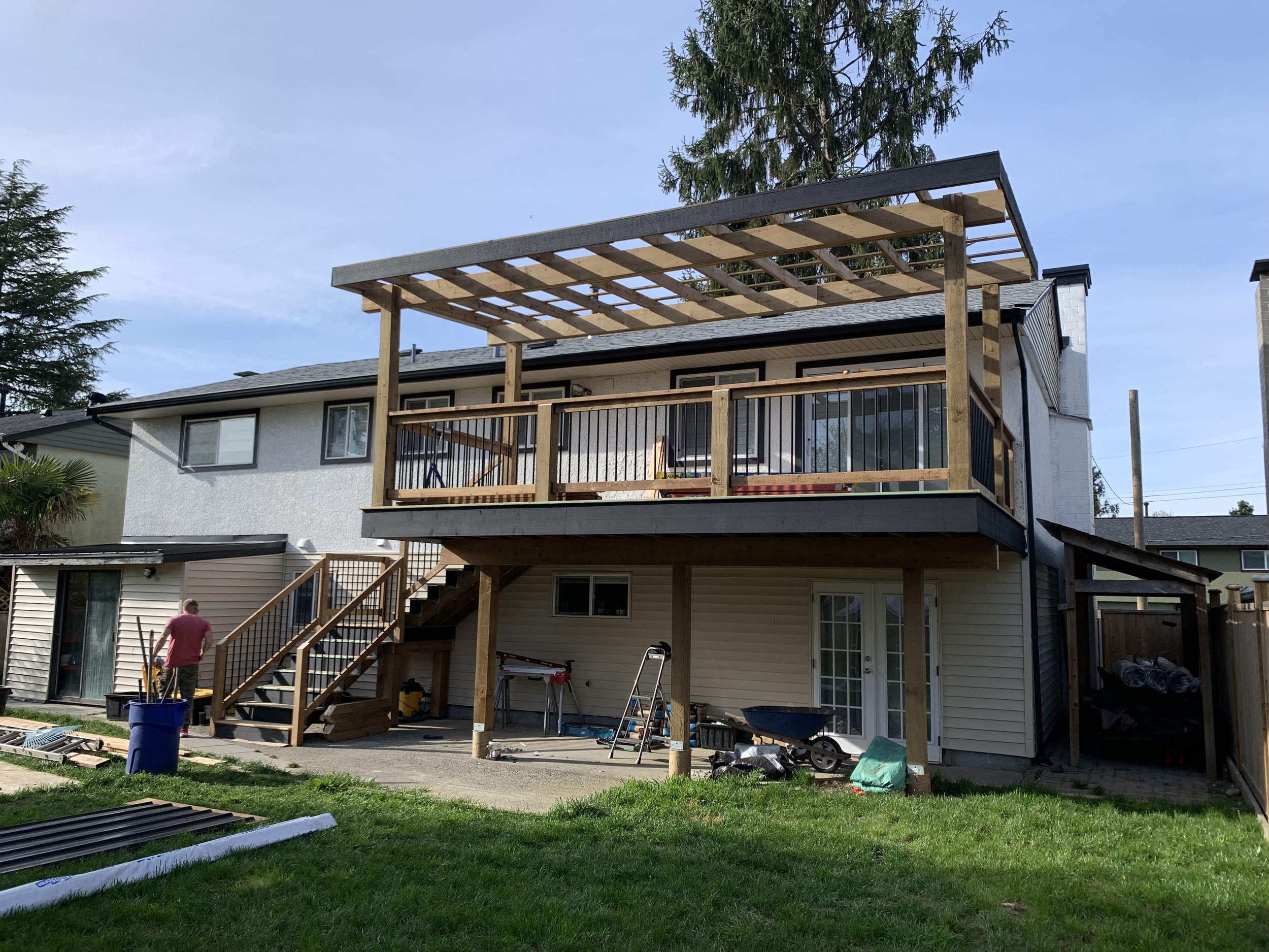 A house with a new wooden deck under construction on the second floor, with tools and workers present, in a suburban backyard with green grass and trees.