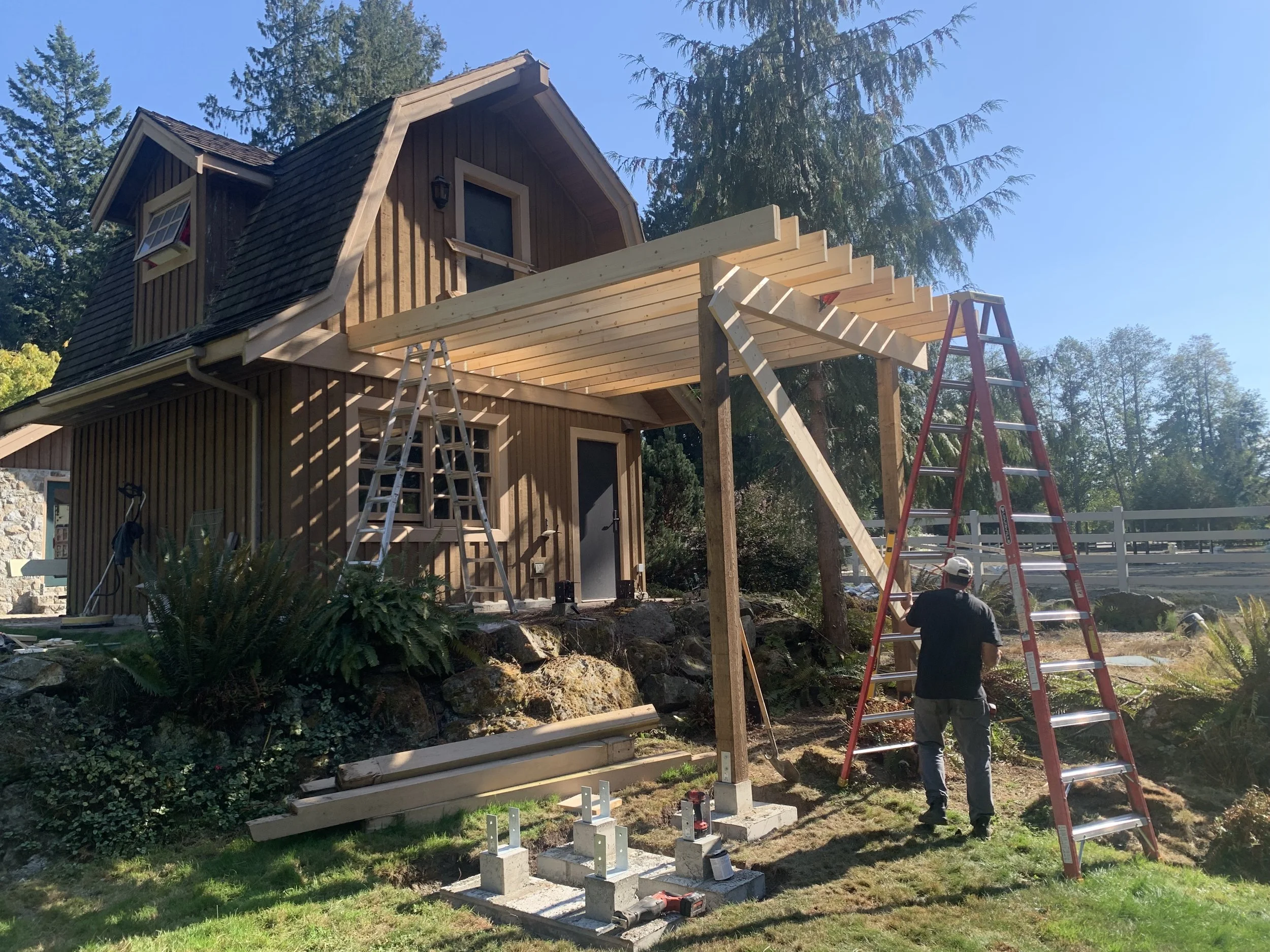 Two men working on construction of a wooden deck attached to a house surrounded by trees. One man is on the ground, and the other is on a red ladder. Various construction materials and tools are visible.