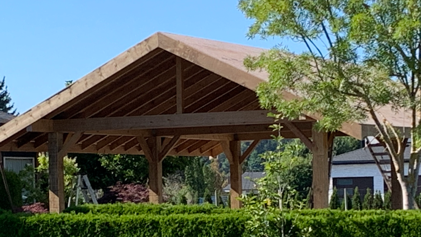 A wooden carport or pavilion with a peaked roof, set in a residential backyard with trees and shrubs, under a clear blue sky.