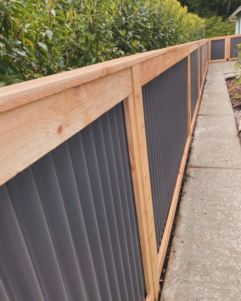 A wooden fence with dark gray corrugated metal panels along a concrete sidewalk, with green bushes and trees in the background.