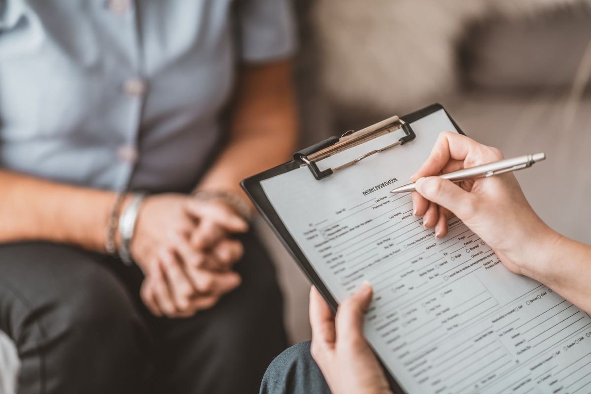Close-up of a healthcare professional filling out a patient registration form on a clipboard, with a patient sitting in front with hands clasped.