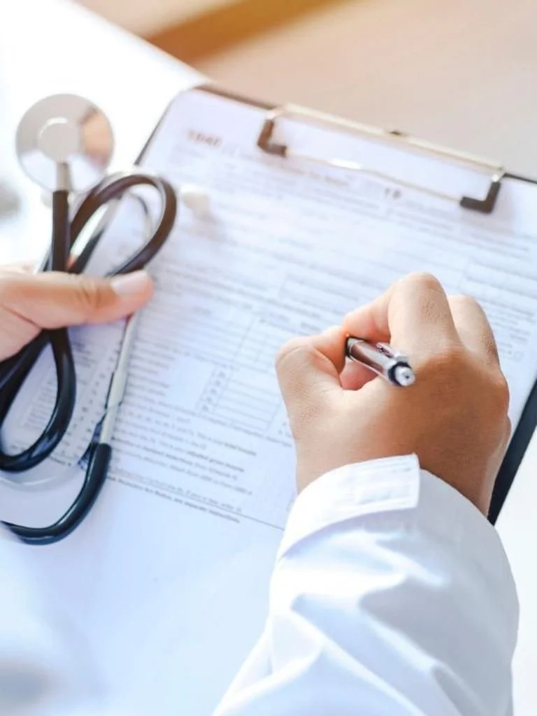 Healthcare professional filling out a medical chart with a pen, holding a stethoscope in their other hand on a clipboard.