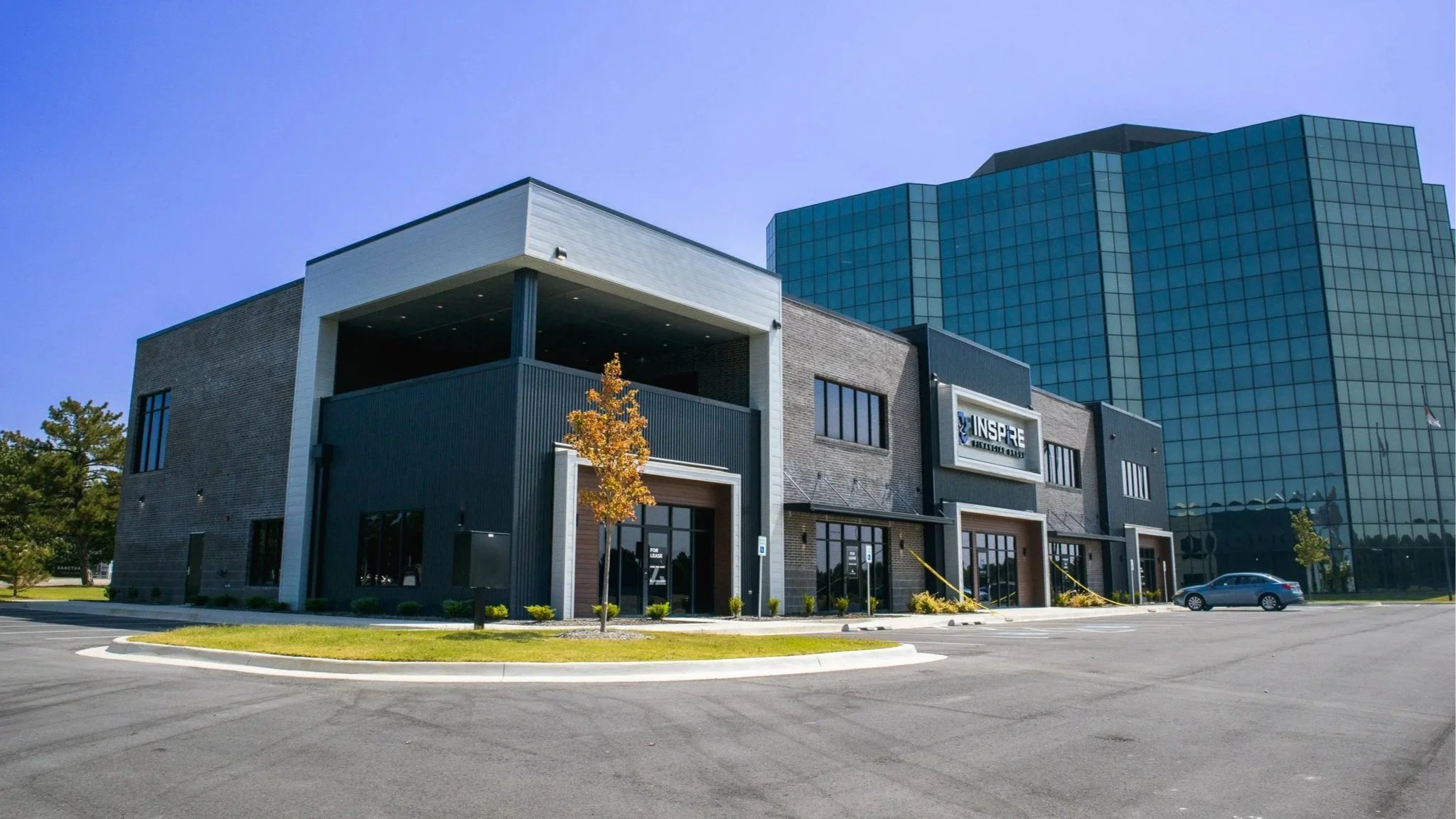 Modern commercial building with brick and glass facade, surrounded by a parking lot with one car and small trees, under a clear blue sky.