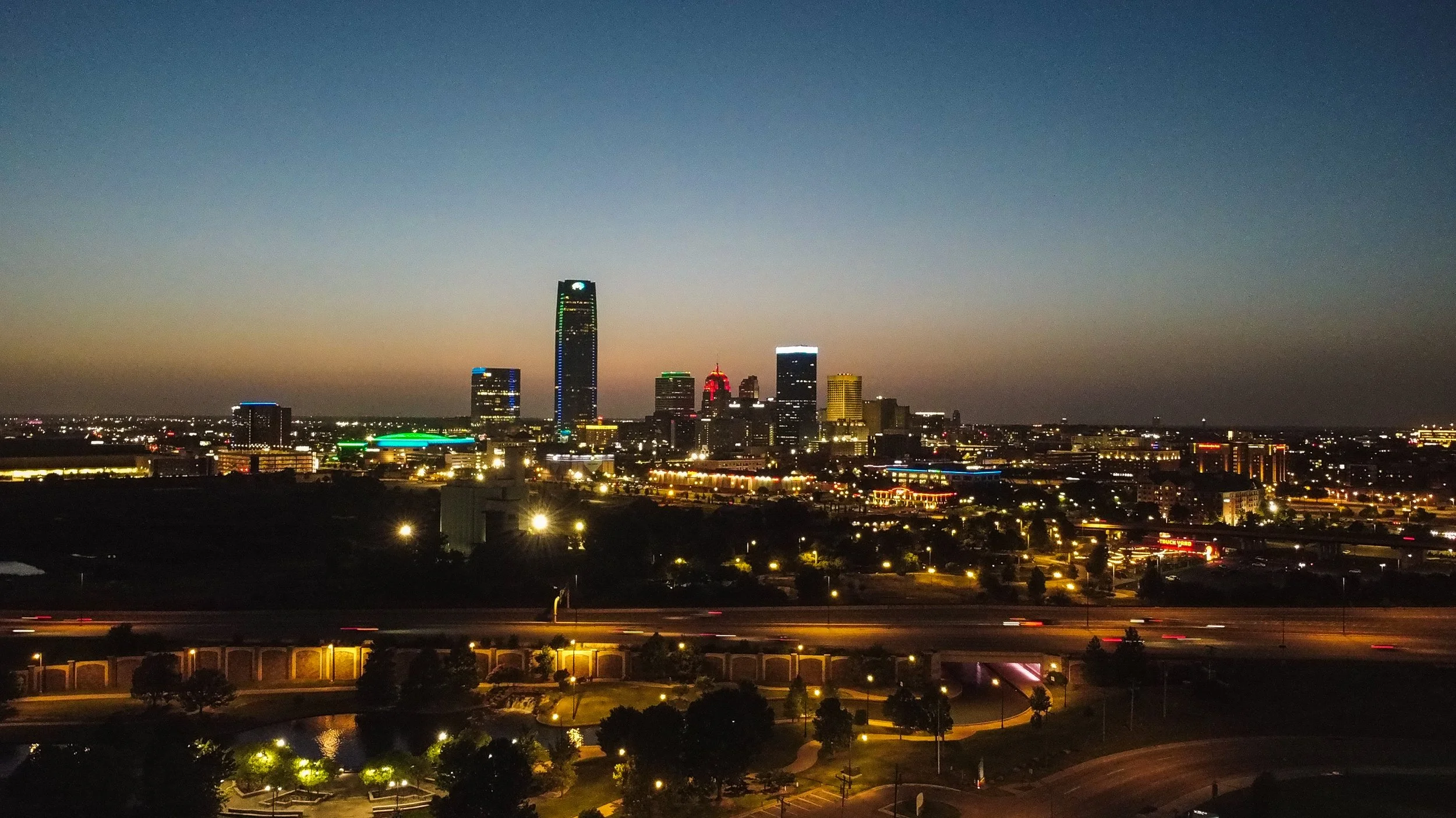Nighttime city skyline with illuminated buildings and a clear sky, including a tall skyscraper, smaller buildings, and city lights reflecting on the ground.