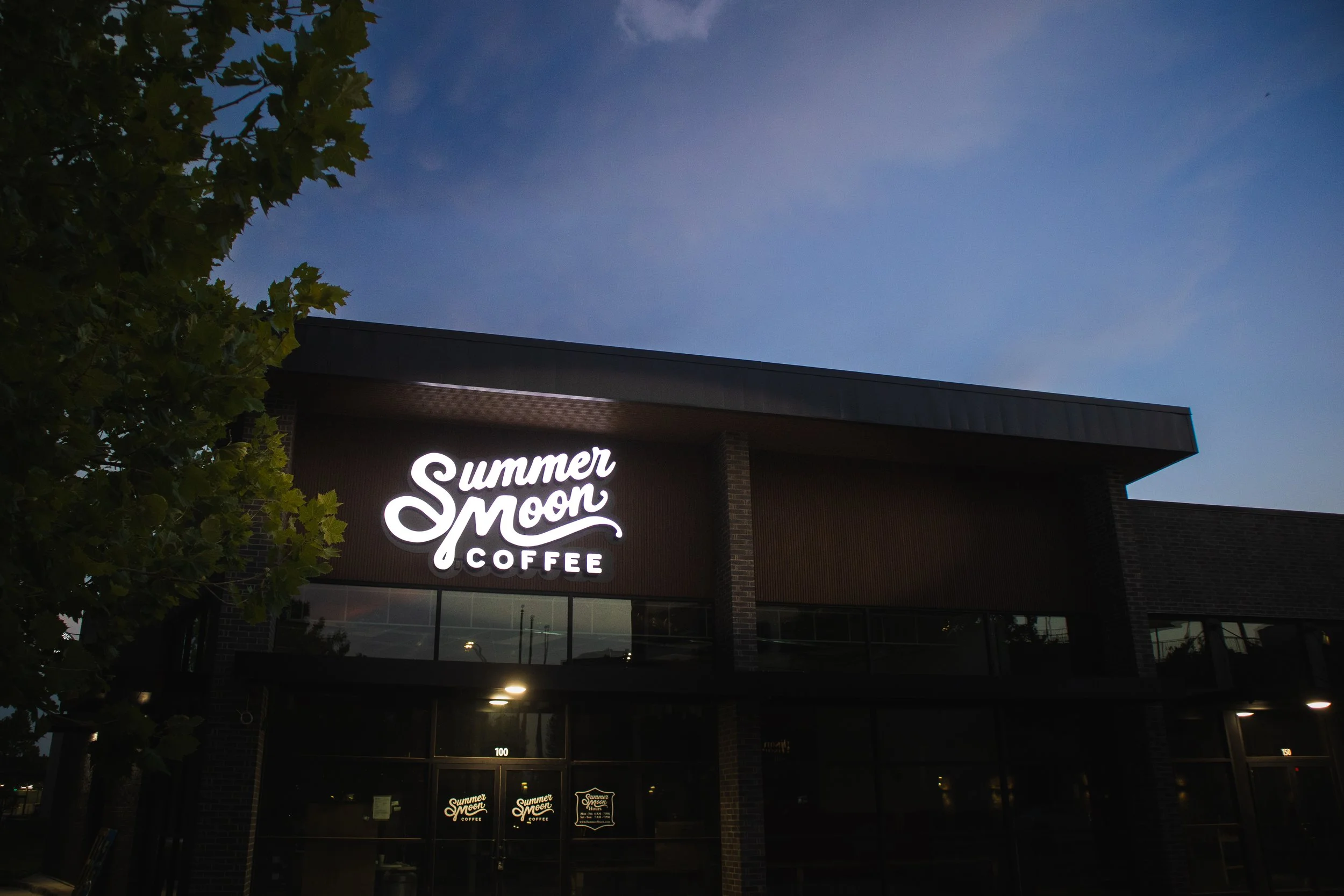 Nighttime view of a coffee shop named Summer Moon Coffee with illuminated sign on the building, partially obscured by tree leaves, and a clear blue sky in the background.