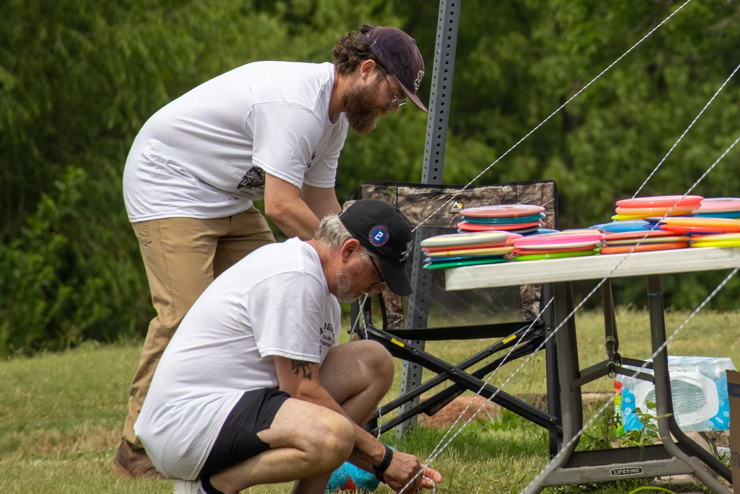 Three men at an outdoor frisbee game setup, with colorful frisbees on a table, and trees in the background.